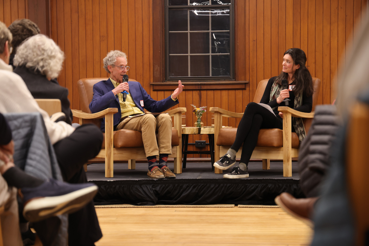 man in sports jacket with microphone sits together with a woman in a chair across from him, both on a raised stage