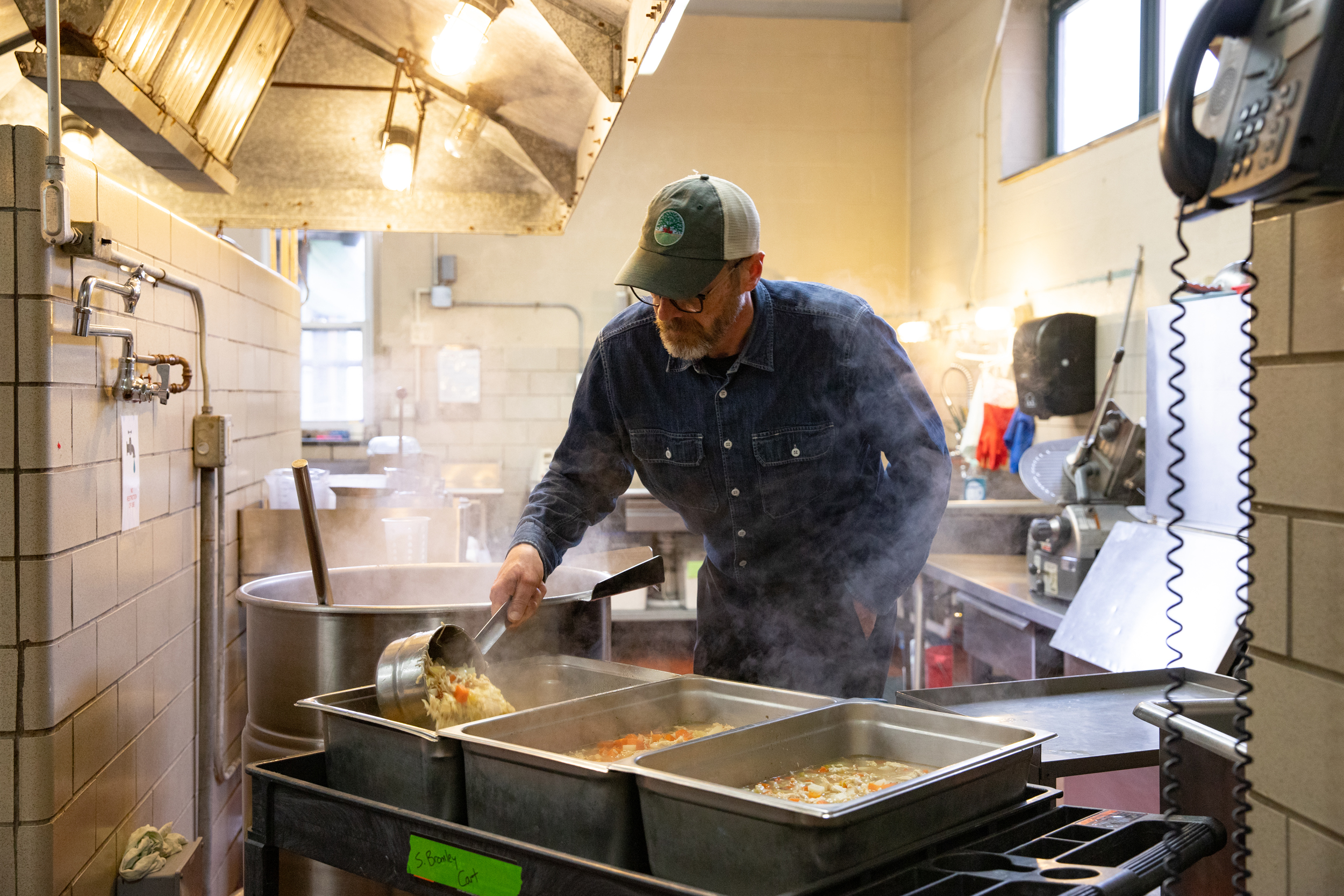 A man ladles soup into large hotel pans in a school kitchen.