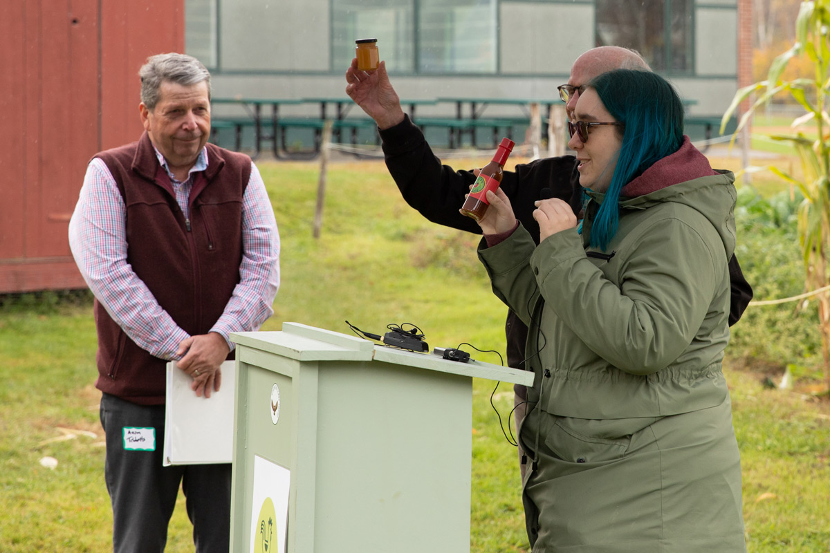 Three individuals at an outdoor event, one standing behind a podium presenting honey and hot sauce as a gift.