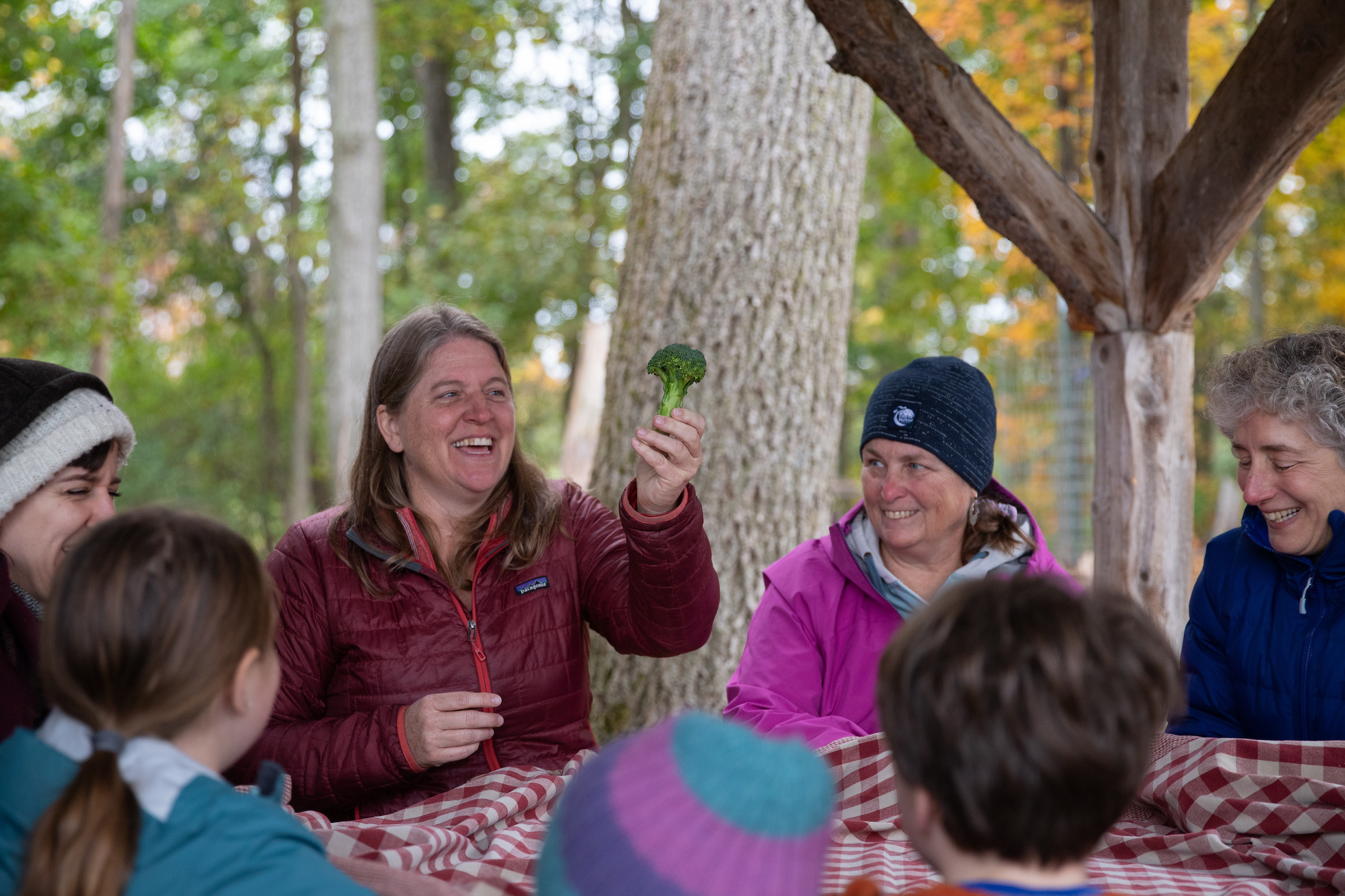 A woman holds up a piece of broccoli, she is part of a group seated in at a picnic table