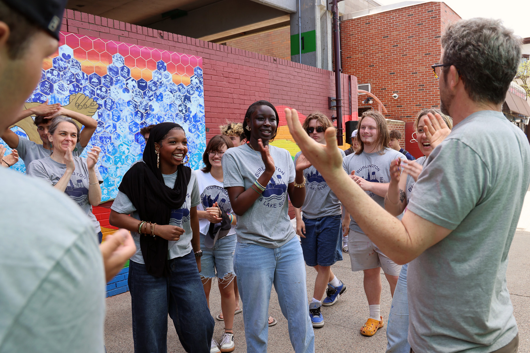 A group of students smile and laugh looking toward an instructor with a colorful mural painted on a brick wall behind them.