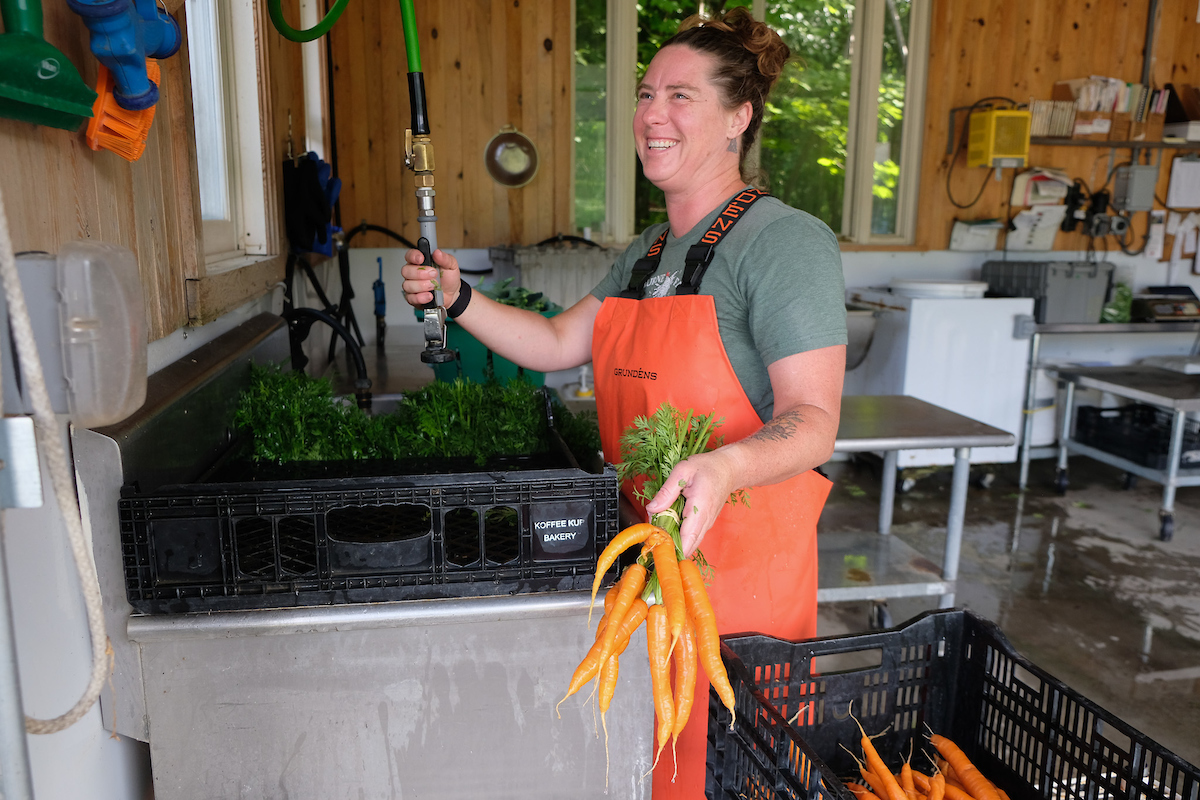 woman wearing waterproof bib laughs as she washes carrots at a basin in an open air wash shed