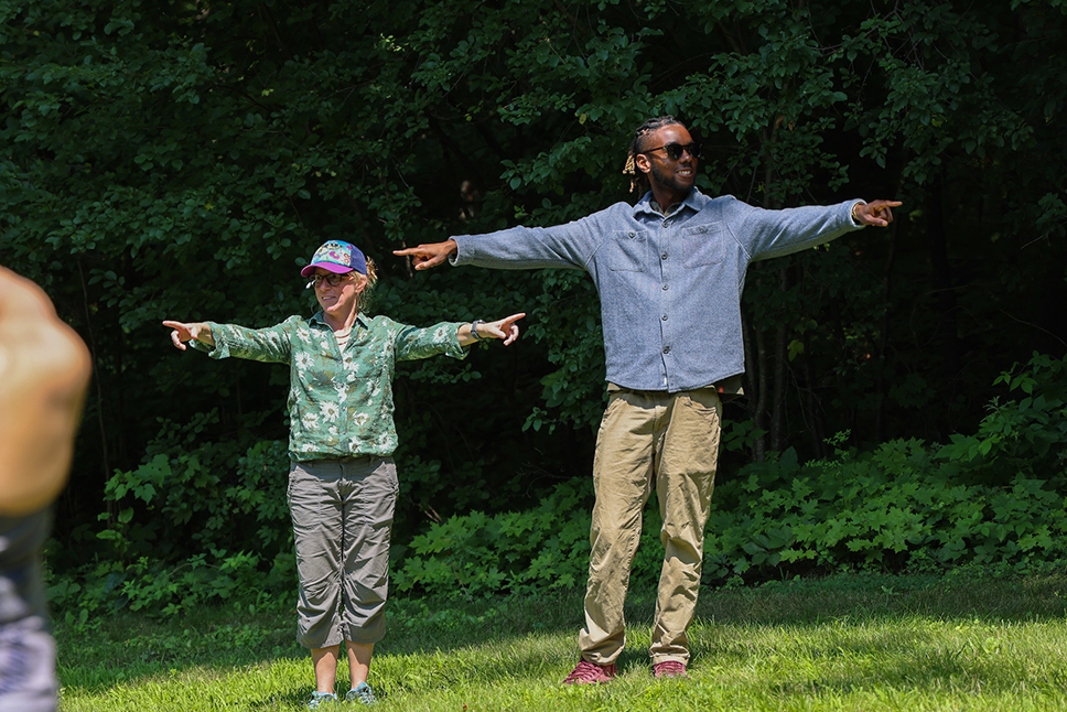 Two adults stand in a grassy, shaded lawn, smiling while pointing to others outside of frame