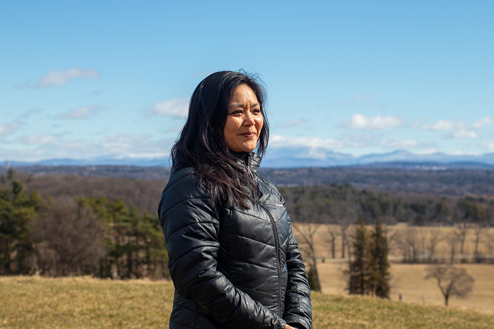 A portrait of Aimee Arandia Ostensen standing on the top of a hill in winter with sweeping views of rolling hills, mountains, and blue sky beyond