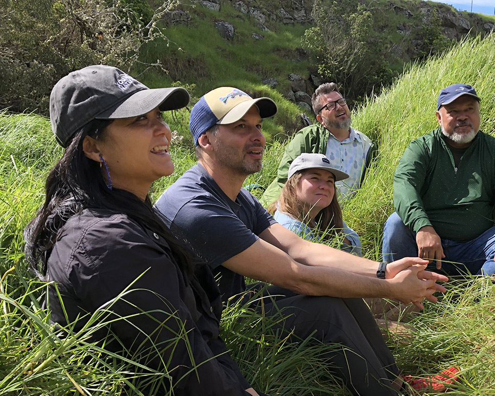 Five educators sit among tall grass, smiling while looking off camera