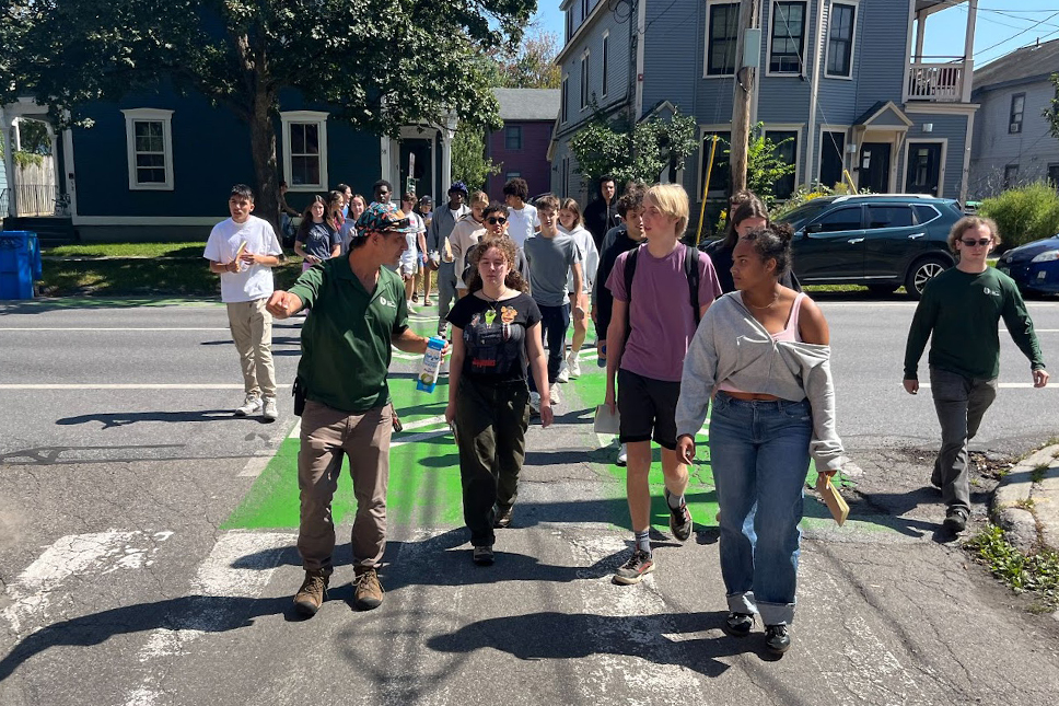 A group of high school aged students cross a paved city street in conversation with each other and an adult tour guide