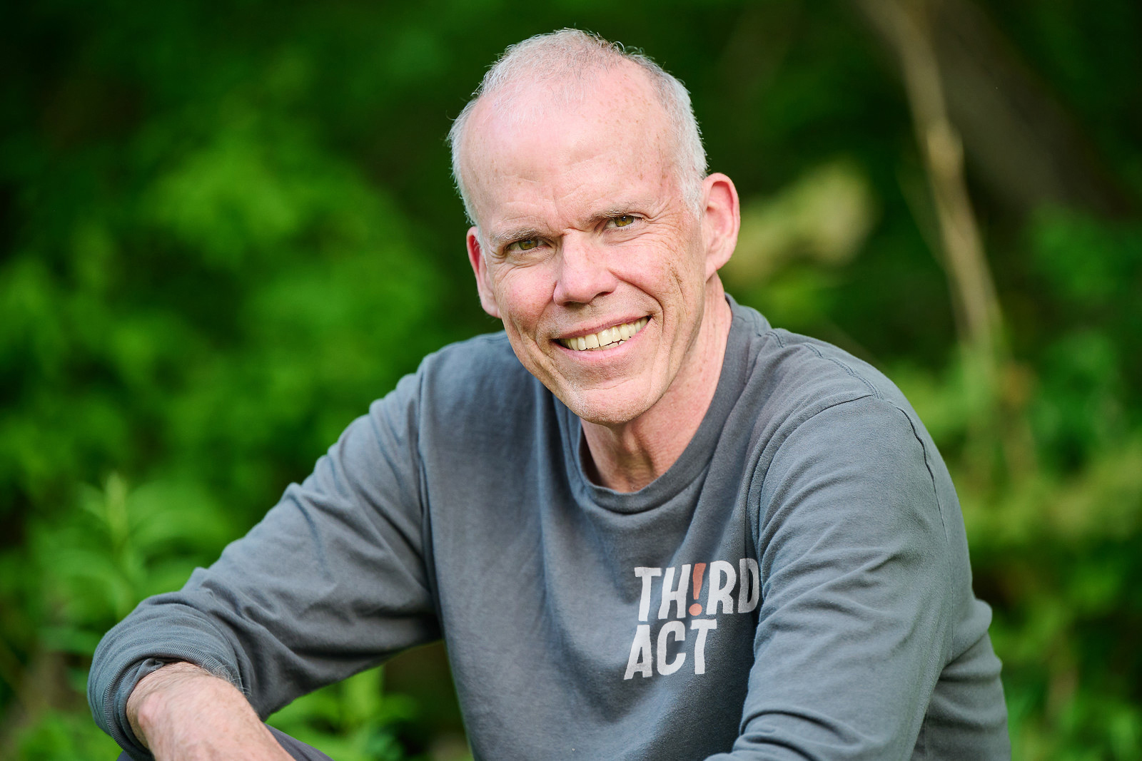 Man with white hair in a t-shirt which reads "TH!RD ACT" smiles in front of a background of green foliage.