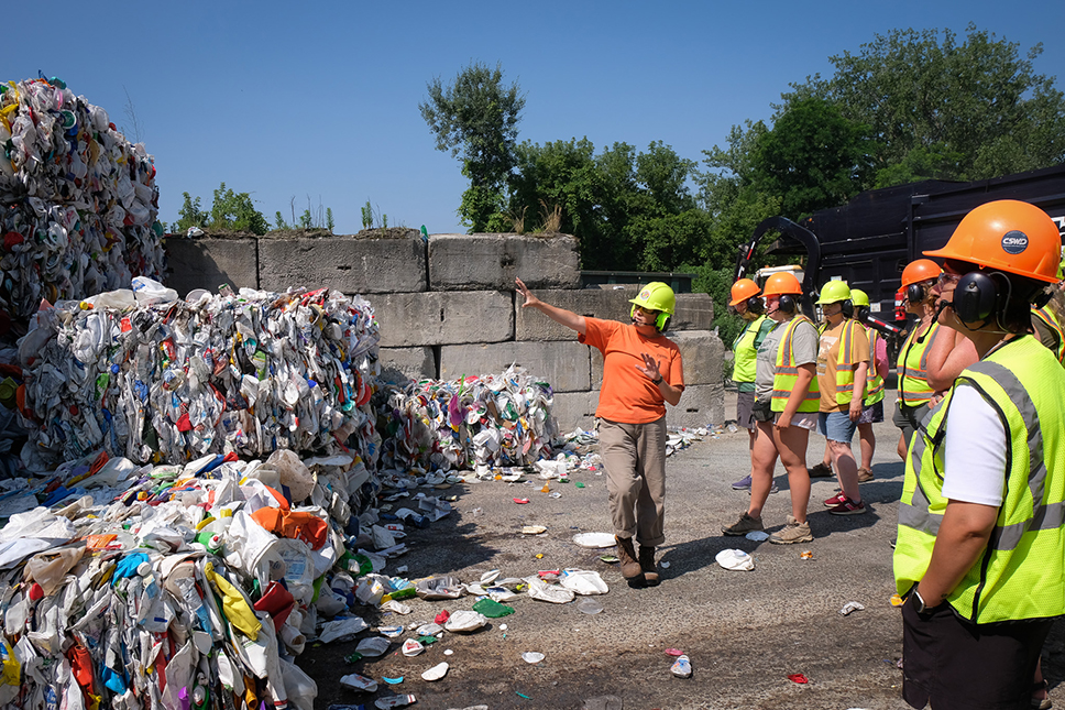 A group of people tour the outdoor area of a recycling facility with large compacted cubes of plastic stacked around them