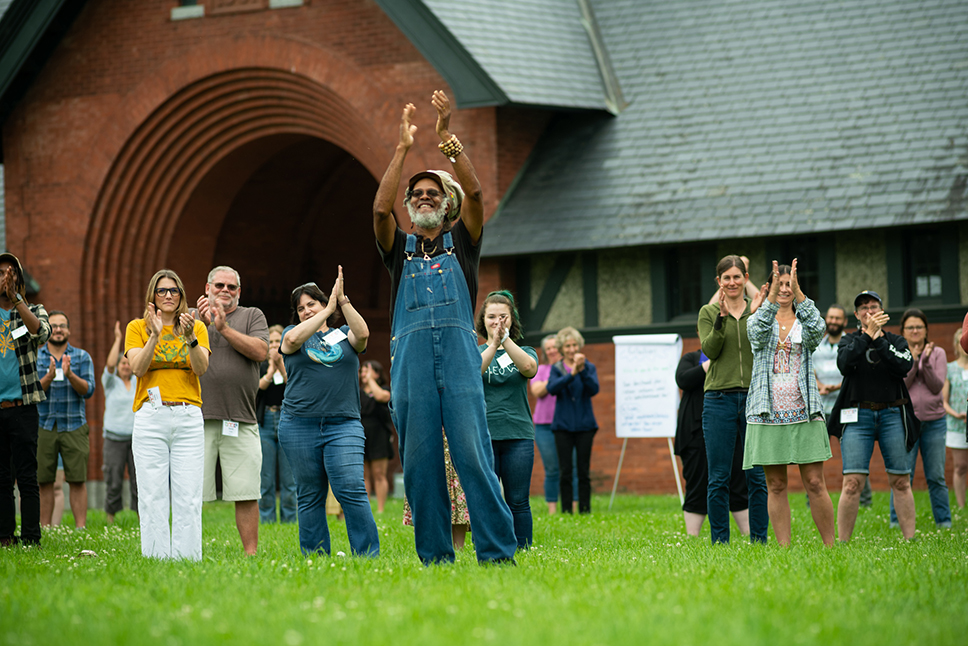 A large group of adults stand in grass smiling and applauding in front of a brick building