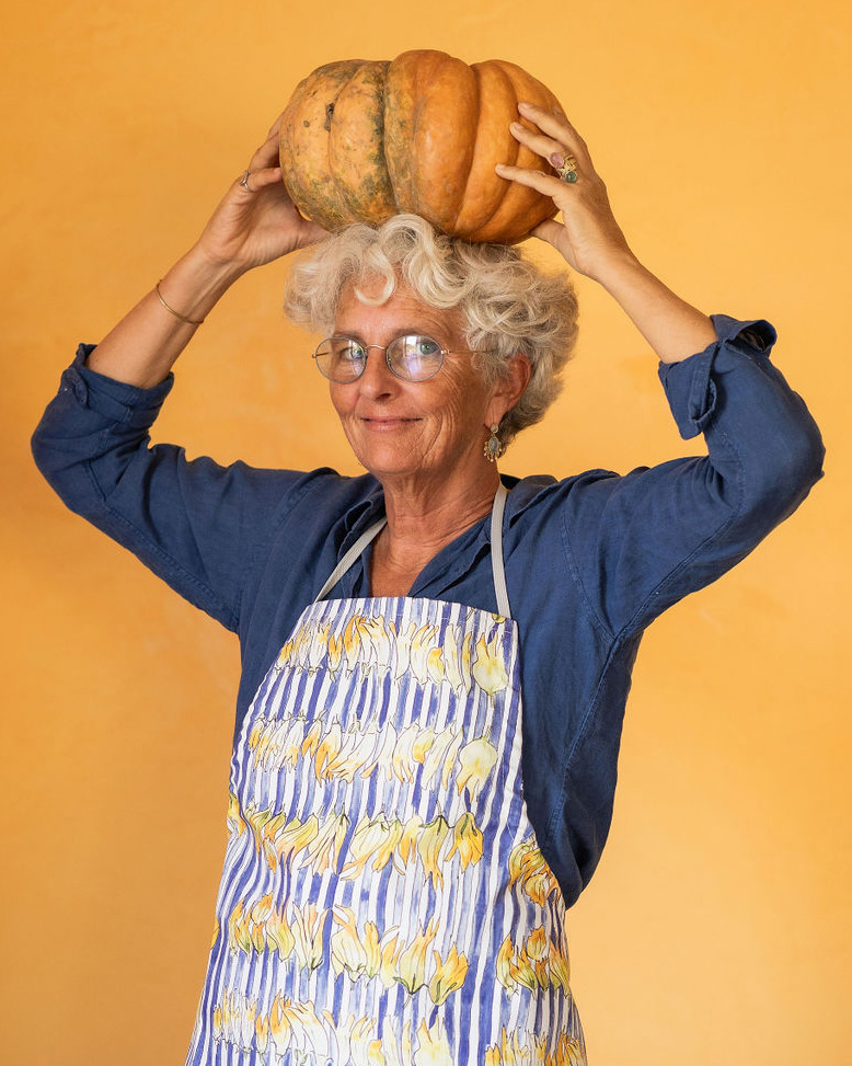 Woman with short gray hair and glasses smiles while holding a pumpkin on top of her head. She is wearing an apron.