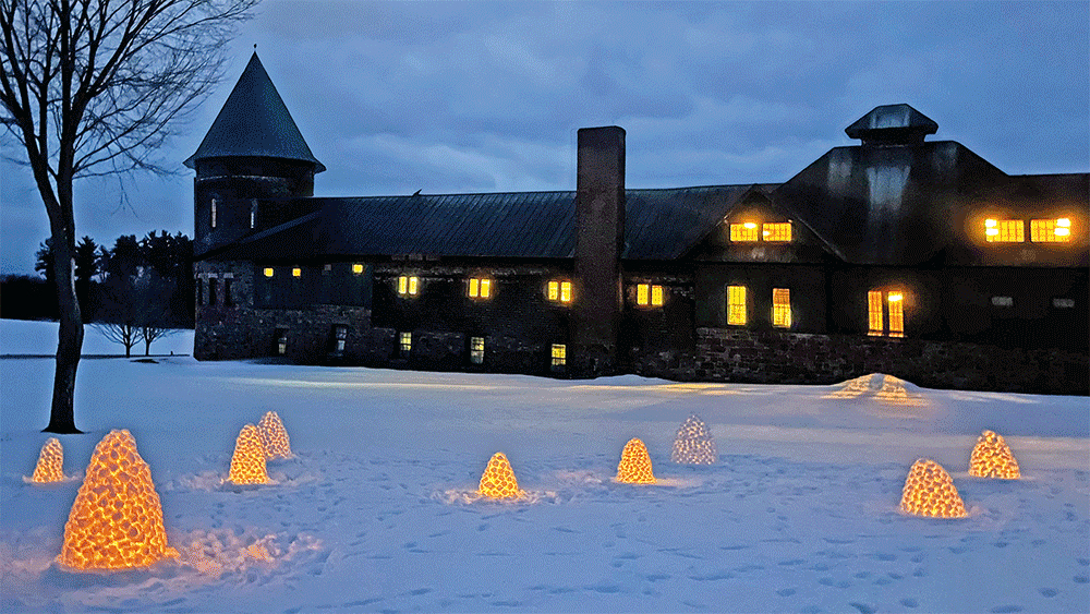 multiple small snow igloos, lit up with candles inside them, in front of the Farm Barn on a winter evening.