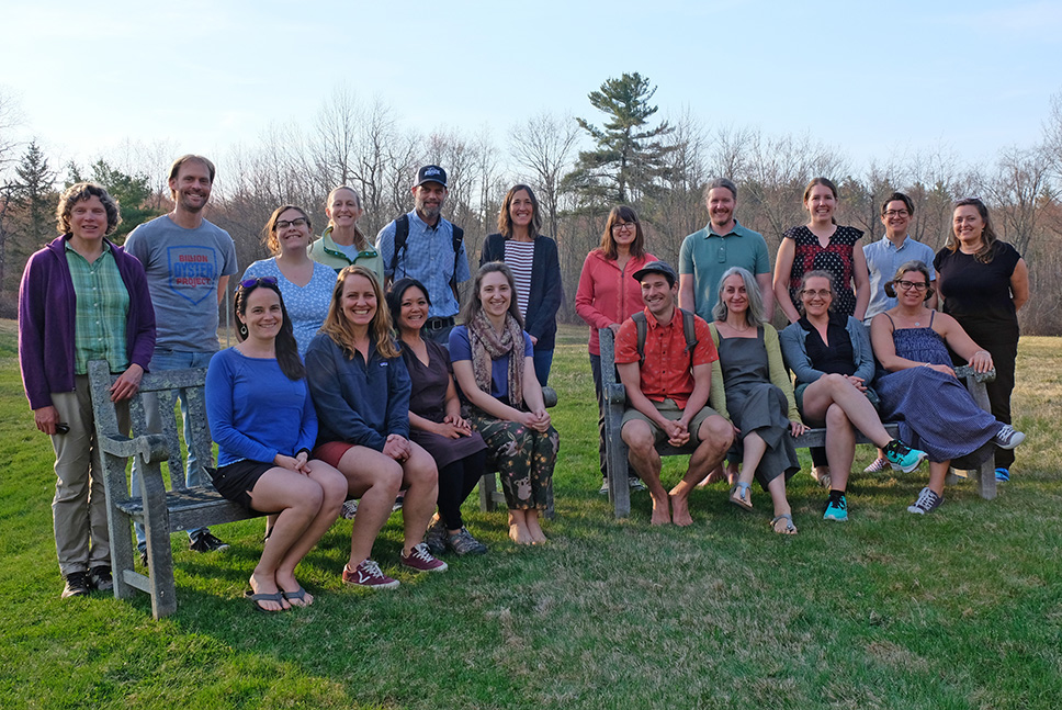 A group of twenty educators pose and smile for camera, seated and standing, at dusk on a grassy lawn