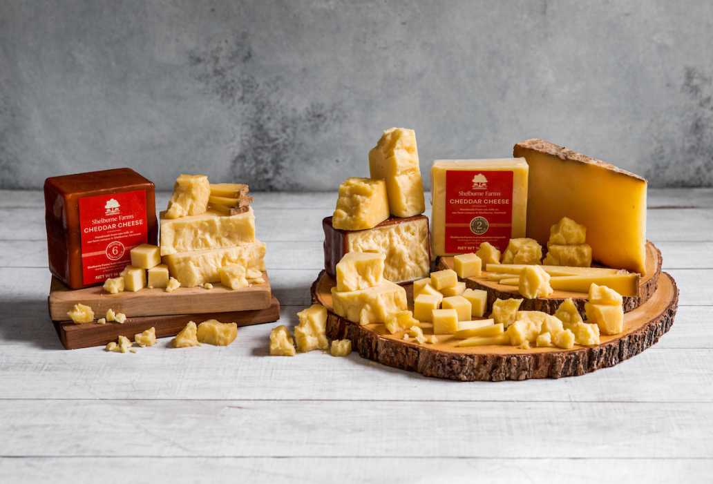 Assorted Shelburne Farms cheddar cheeses displayed on wooden boards. Some pieces are crumbled around the boards for a rustic look on a grey background.