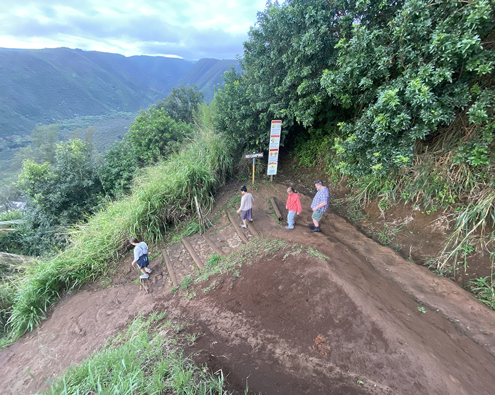 Four adults hike down a red dirt switchbacking trail, with Hawaiian landscape of mountains and clouds in distance