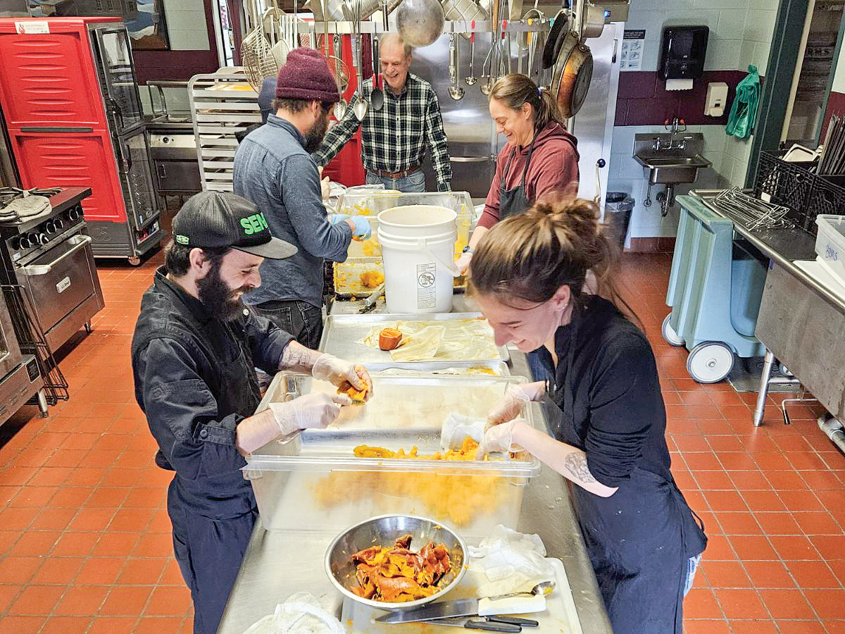 A group of people works together in a school kitchen to process squash around two stainless steel tables.