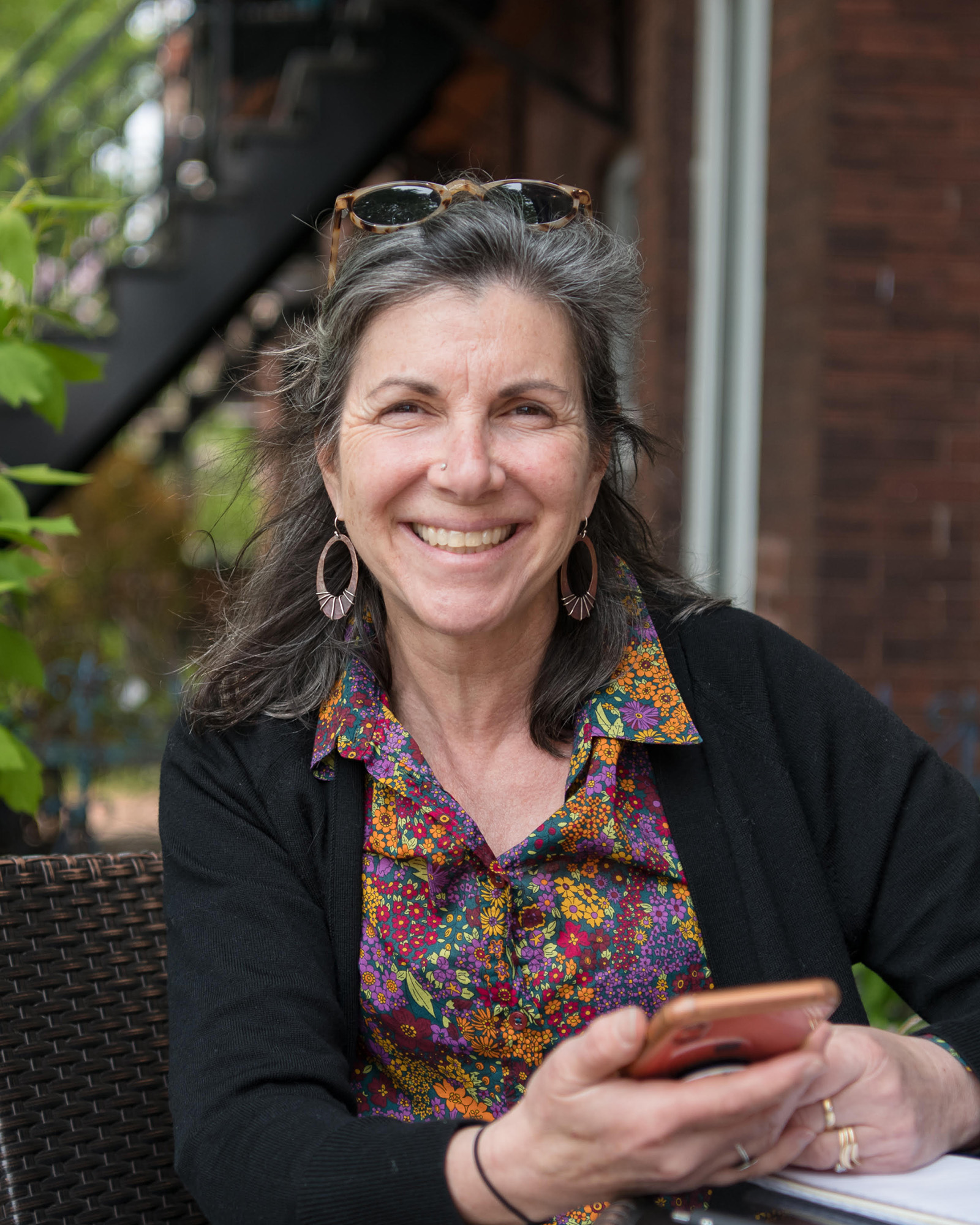 Woman with gray hair smiles at camera in an outdoor setting with a brick building and green foliage behind her.