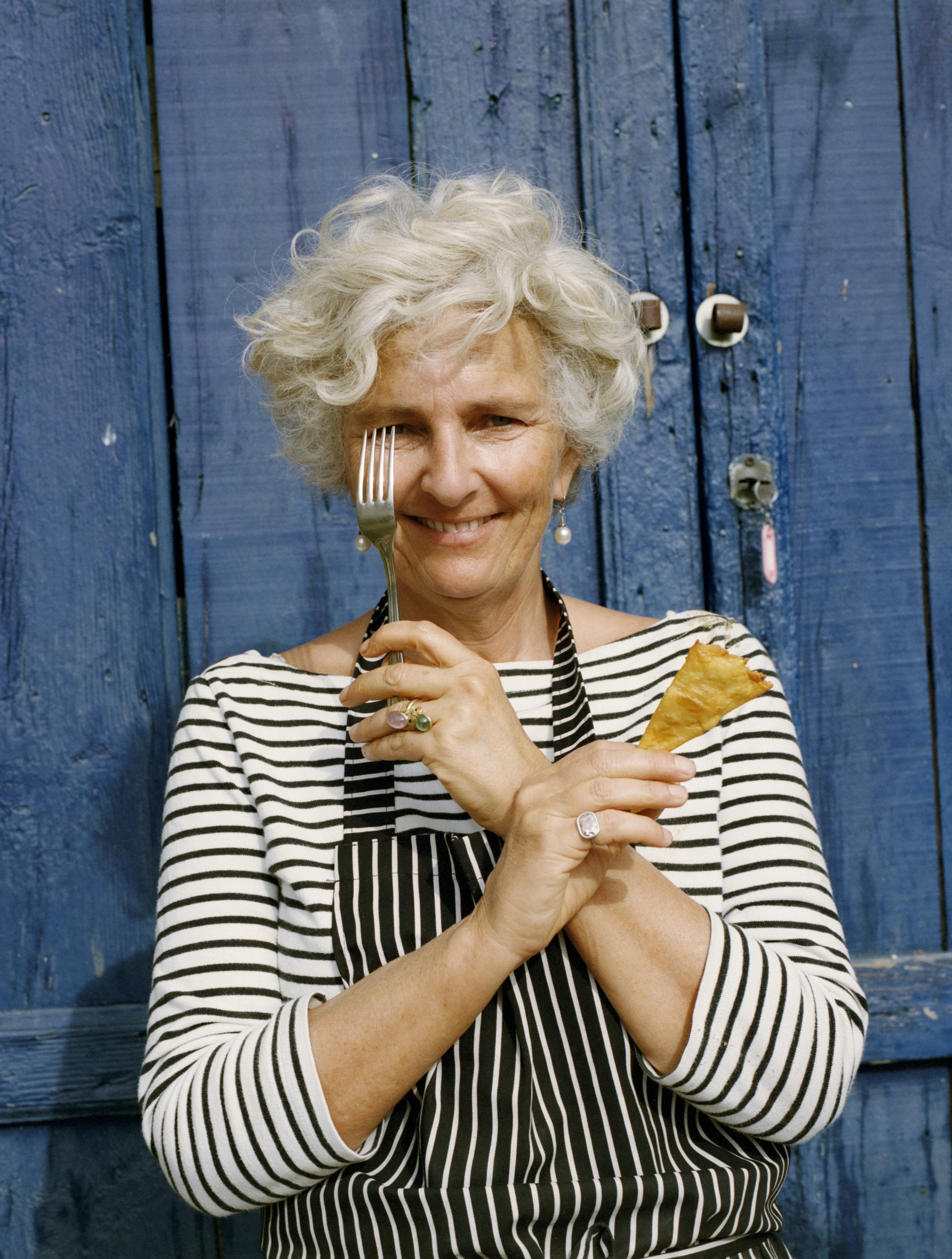 Woman smiling holding a fork over her eye and a piece of food in her other hand in front of a blue wall.