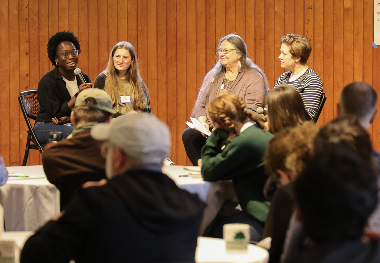 Four women sit on a stage in a panel discussion in a wood paneled room