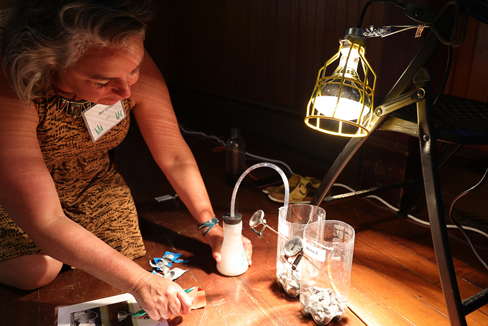 A woman kneels on a hardwood floor in front of a science experiment involving two plastic bottles and a lamp