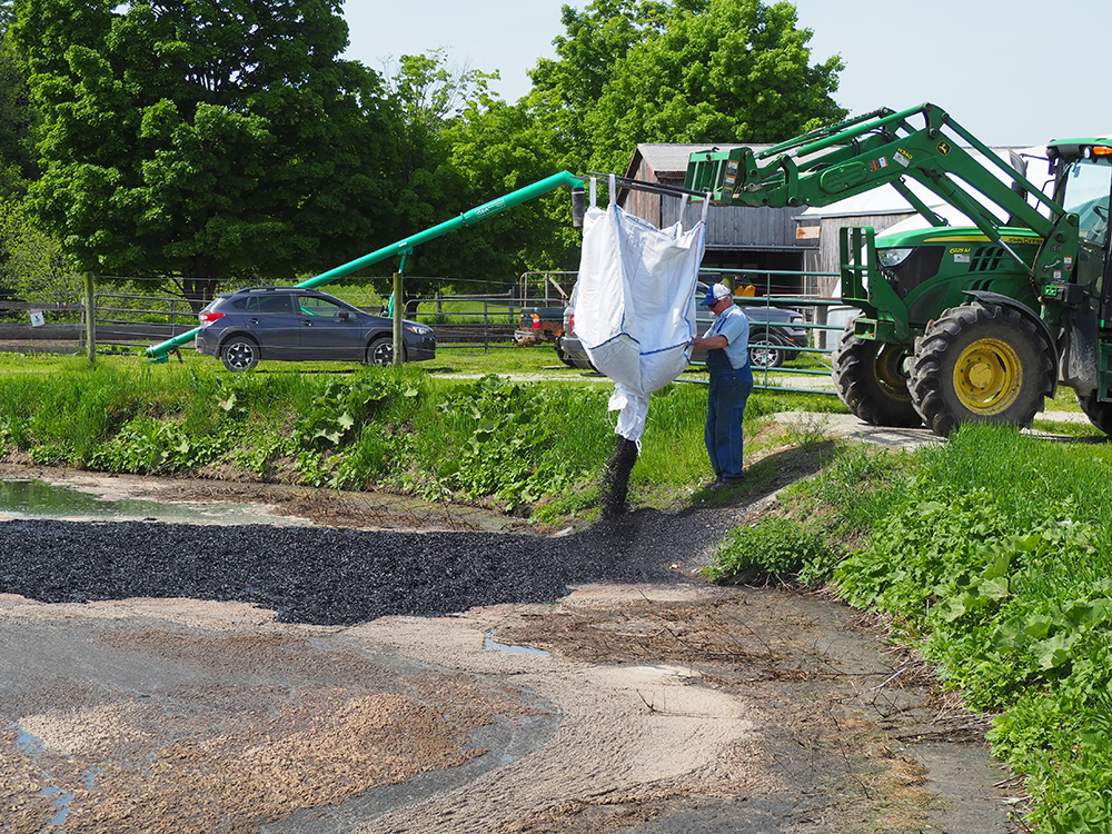 man in overalls guiding big white bag of biochar hanging from a fork lift as it spills into a manure pit.