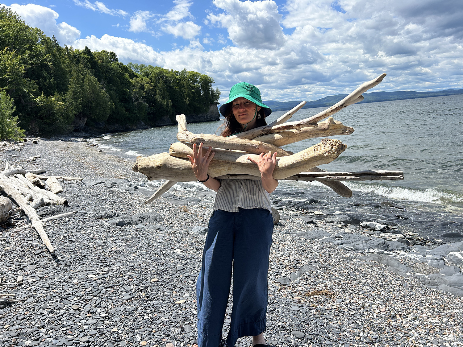 woman in a sunhat standing on a rocky beach holding an armful of driftwood on a sunny day