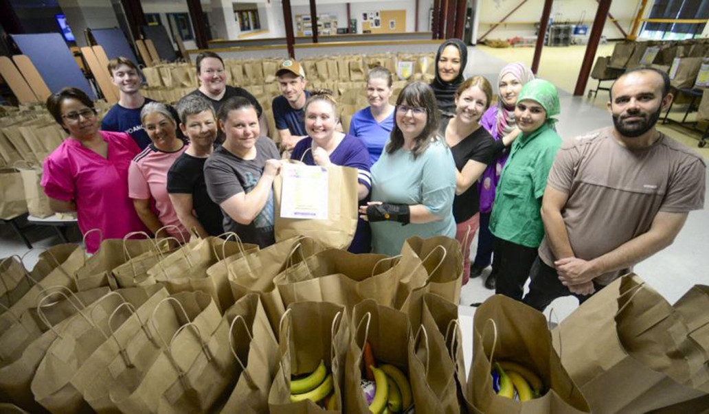 Group of school meals staff at a school cafeteria, standing behind rows of filled grocery bags.