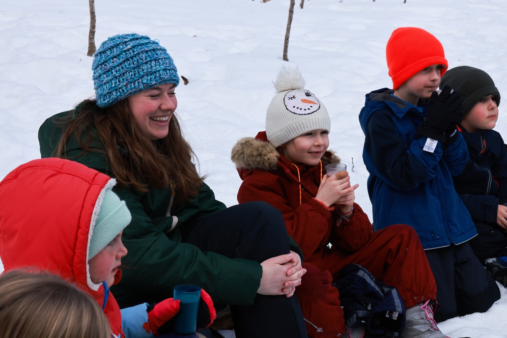 Multiple children and one adult, all in winter clothing, sit and smile surrounded by snow.