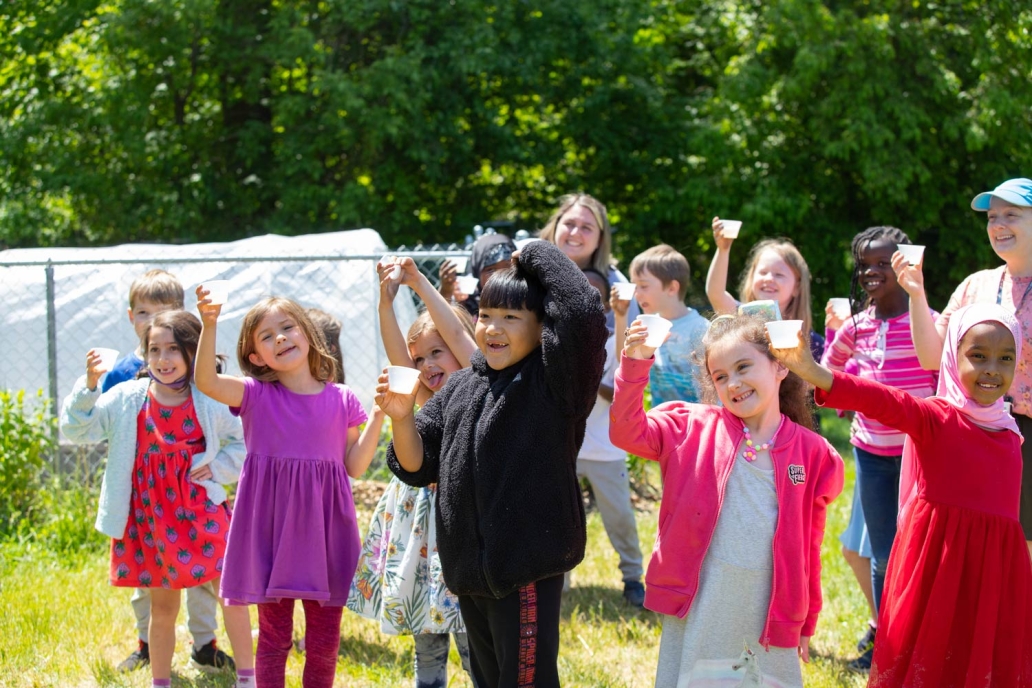 A group of smiling children holding up cups in a sunny outdoor setting, with greenery and a fence in the background.