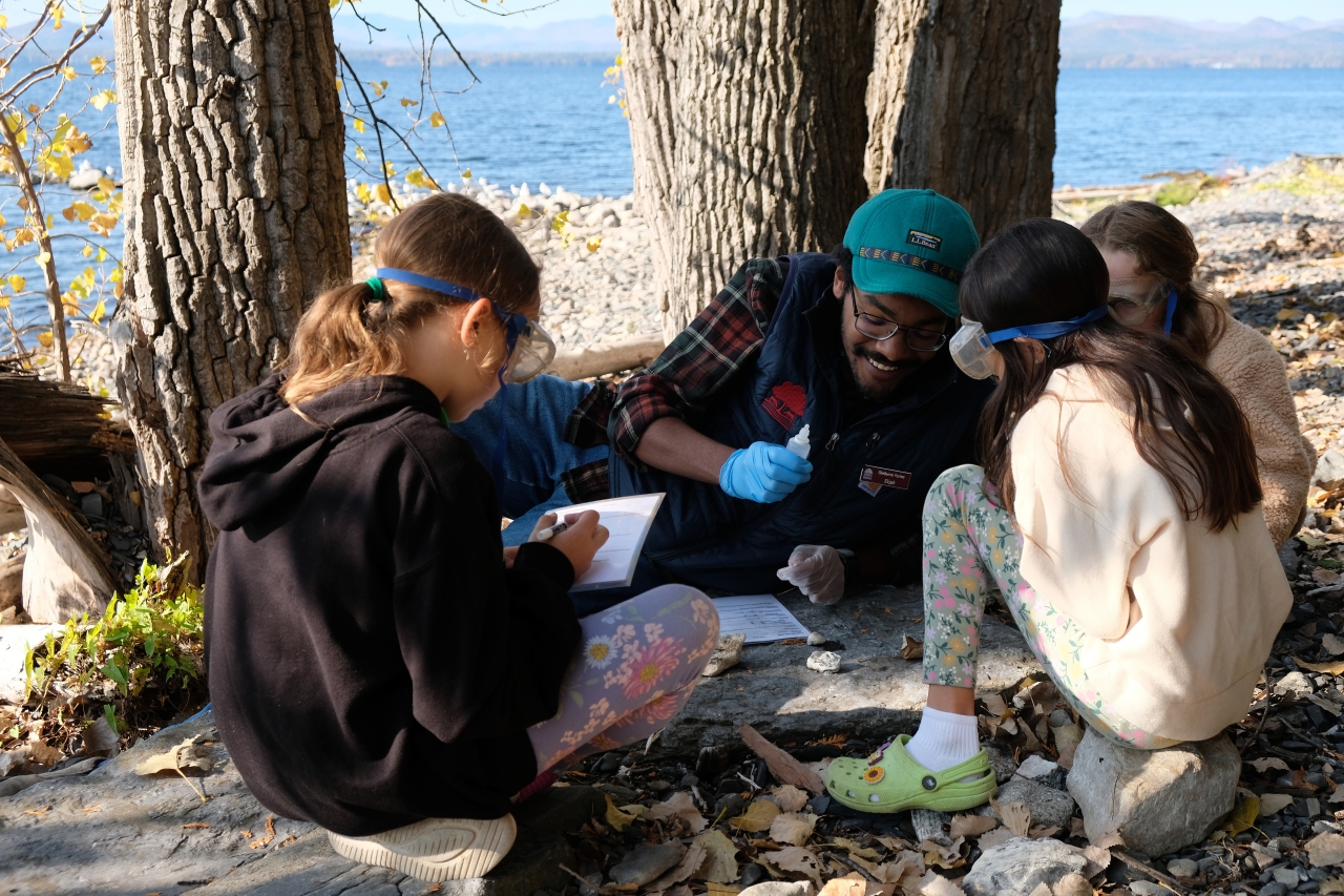 Three young children gather around an educator conducting an experiment with rocks on a rocky lakeshore.