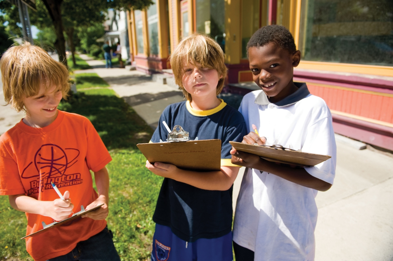 Three young children hold clipboards and smile while standing on a city sidewalk