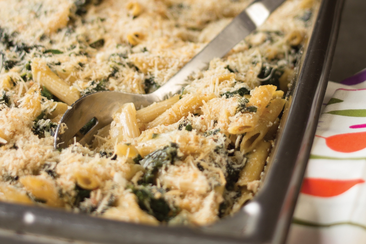 Baked pasta with kale and cheese in a glass dish, served with a spoon.