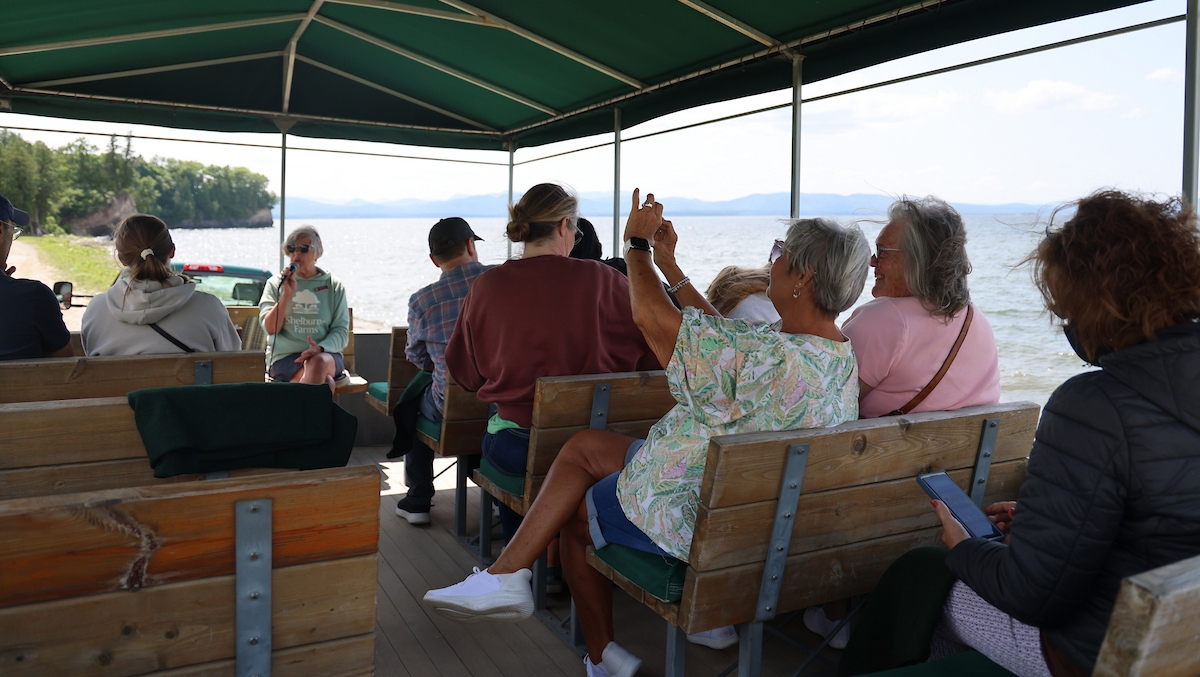 group of people on a open-air wagon facing tour guide at the front with lake in the background