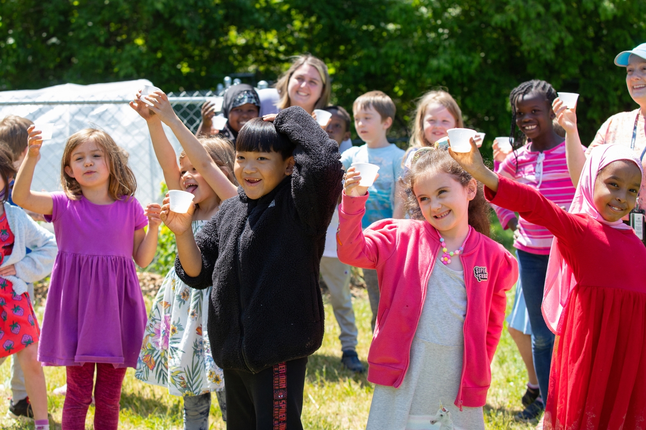 A group of young children smile while holding up paper cups in a garden