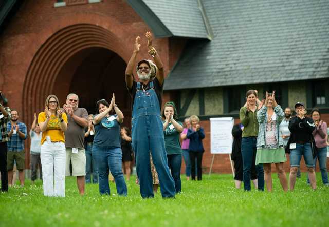 A large group of adults stand in grass smiling and applauding in front of a large brick building