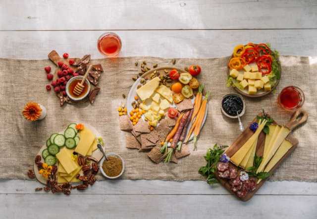 An assortment of cheeses, meats, and vegetables neatly arranged on a rustic wooden table, accompanied by condiments and drinks. Decorative flowers add a touch of color to the setting.