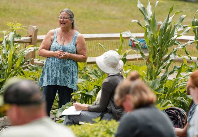 A woman stands in a garden with raised beds full of corn squash and beans, smiling while talking to a group of seated people