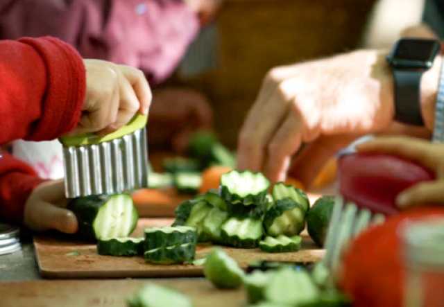 Close-up of hands slicing cucumbers on a wooden cutting board at a brightly lit table.