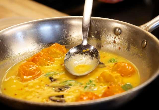 Close-up of a stainless steel skillet containing a sauté of cherry tomatoes and herbs in a yellow sauce, with a spoon resting inside.