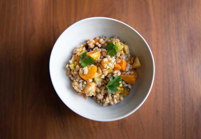 A bowl of wheat berries with roasted squash and parsley garnish, on a wooden table.