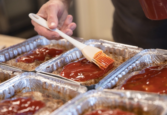A person applies barbecue sauce to meatloaf using a brush in aluminum trays on a kitchen counter.