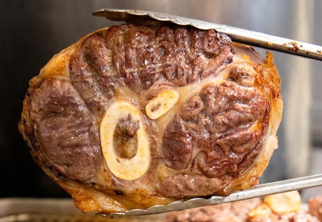  Close-up view of a roasted beef shank with a bone marrow section, glistening as it is held up by tongs.