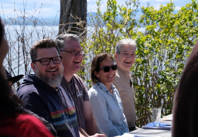Four educators laugh around a sunny, spring picnic table at lakeshore at Shelburne Farms. Green bushes stand in background with lake and Adirondack Mountains in distance