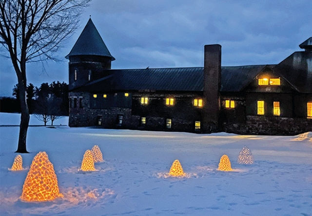 farm barn at night with lit windows and snow candles lit on snowy field in foreground