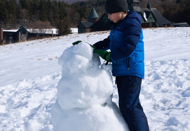 A boy builds a snowman on a snowy field with sweeping views of a large barn and mountains in distance
