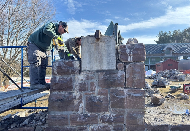 Man on scaffolding with power tool dislodging a large granite block from a half-dismantled pillar of redstone