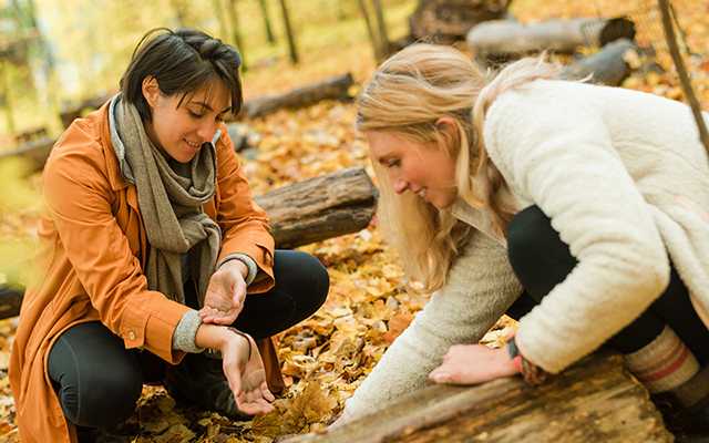 Two people smile while kneeling on the forest floor in autumn, looking for life under a log