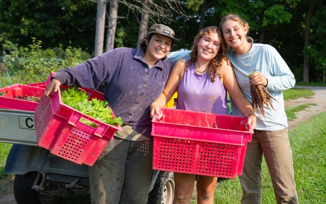 Three farmers smiling at the camera, each holding a red crate filled with fresh vegetables. They are standing outdoors with greenery in the background.