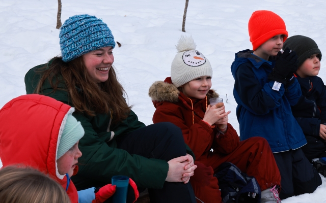 Multiple children and one adult, all in winter clothing, sit and smile surrounded by snow.
