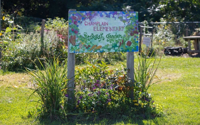 A hand painted sign with "Champlain Elementary Fresh Garden". A garden is in the background.