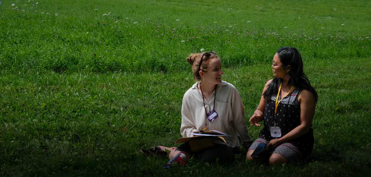 Two people kneel in a sunny grassy field smiling and talking while holding notebooks and papers